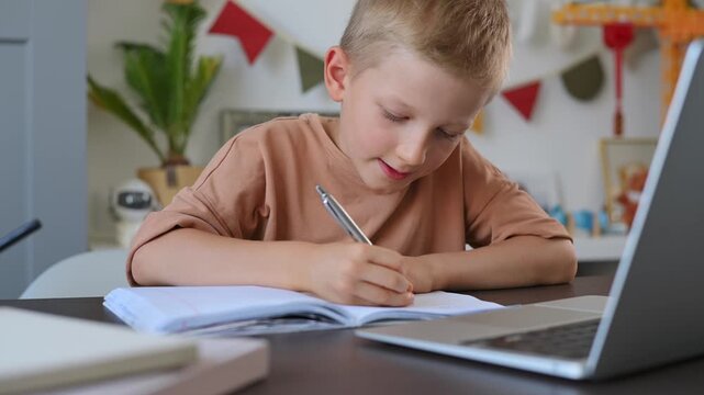 Blond boy writes in notebook and looks at laptop at desk in his child's room. Modern online learning for children, homework done via video link with teacher. Schoolchild studies using laptop. 4k