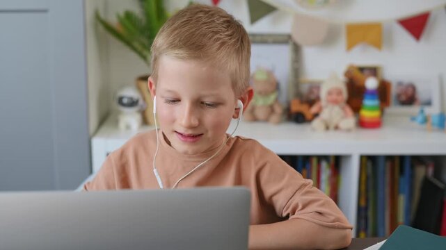 Small fair-haired boy wearing headphones is video chatting with his teacher. Schoolboy is smiling, looking at his laptop, and talking to tutor at his desk. Online learning, video chat, e-lesson. 4k.