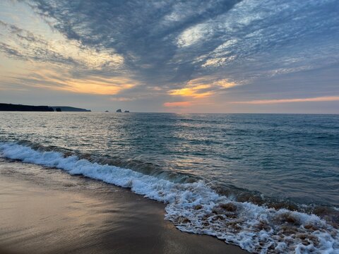 Textured gray and golden clouds sweep across the twilight sky above the Atlantic as a wave meets the sandy shore. Silvery water reflects the fading light in a dramatic coastal scene.