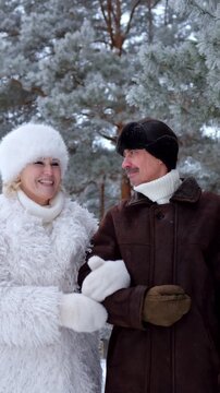 Cheerful elderly couple walking together and lovingly looking at each other in a beautiful snowy pine forest, enjoying their romantic winter vacation in warm clothes and happy mood
