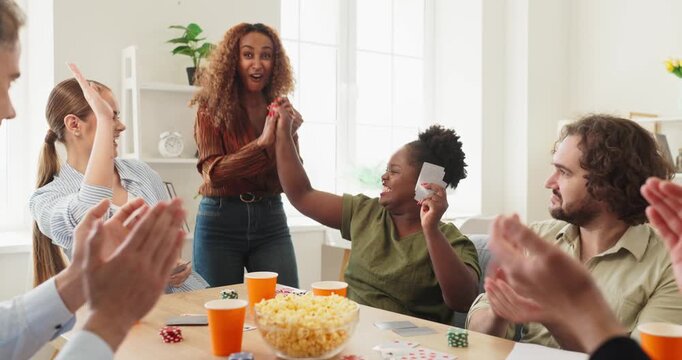Friends players cards poker game cheering winners. A diverse group celebrates a winning hand at home, clapping and laughing. Ideal for social ads and leisure. Concept is teamwork and celebration.
