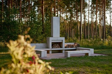 Fototapeta premium An outdoor fireplace seating area at the edge of pine forest, presented with stacked firewood under warm afternoon side light.