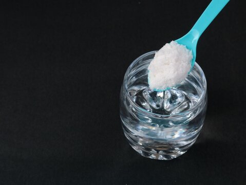 A glass of saltwater solution with blue spoon filled with sea salt crystals on a dark background, representing a saline solution, home remedy, or natural health treatment concept