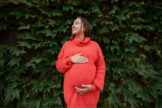 Young pregnant woman wearing red knitted dress and touching stomach in front of plants
