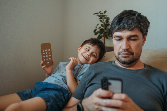 Father works on smartphone while son plays with cardboard toy phone