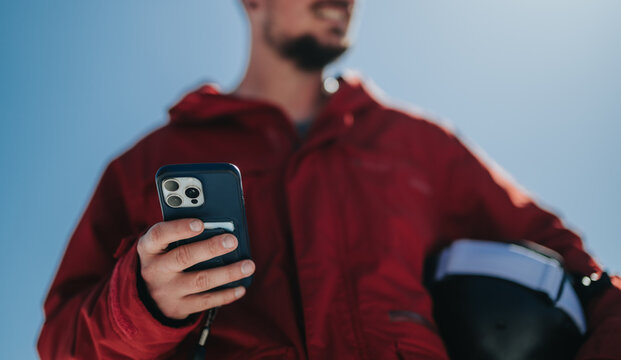 A man wearing a red jacket holds a smartphone while carrying a motorcycle helmet. The outdoor scene shows casual technology use and protective gear in bright sunlight.