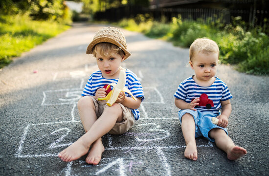 Portrait of toddler boy and his little sister sitting side by side on the street eating bananas