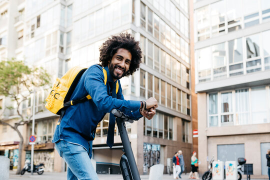 Portrait of smiling young man with yellow backpack on E-Scooter in the city, Barcelona, Spain