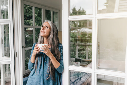 Smiling woman leaning at door frame of her winter garden with cup of coffee looking up