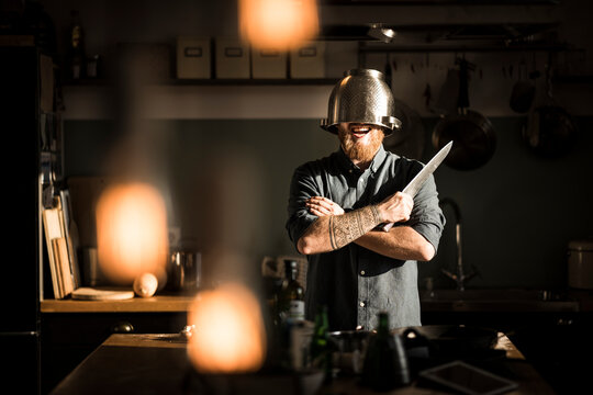 Man with kitchen knife standing in kitchen, wearing colander as helmet