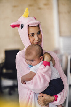 Young mother wearing unicorn onesie, standing in office, holding her son in her arms