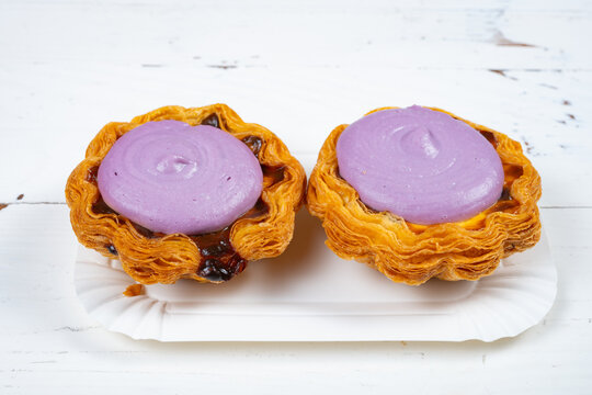 Two flaky egg tarts topped with purple cream on white paper plate