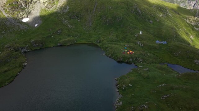 Forward descending drone flight toward a mountain lake surrounded by alpine peaks, with tents and visitors gathered near the water and a small patch of lingering snow visible in the landscape.