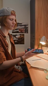 Vertical of Caucasian teenager sitting at desk, picking up smartphone from stand and scrolling through screen. Footage suitable for mobile app marketing, digital lifestyle, or elearning campaigns