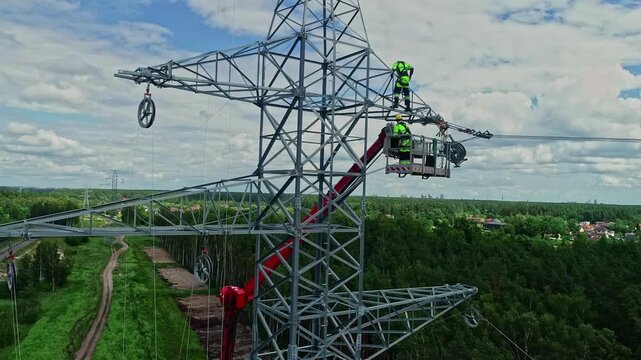 Latvia - 18 February 2023: Aerial view of technicians working on a high voltage electricity pylon, with a bucket truck and lush forest landscape in the background.