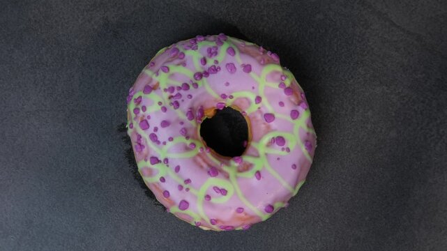 Colorful donut with pink icing and green drizzle on a dark surface, top view, donut spinning