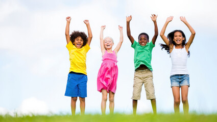 Fototapeta premium Four children jumping with arms raised on grass, showing joy and energy with a bright sky background. Suitable for family, education, and health themes with copy space.