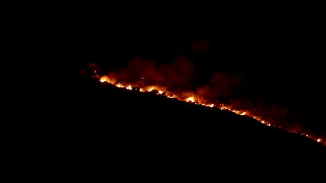 A long line of bright orange fire burns aggressively along a dark mountain ridge at night. This tragic environmental disaster illustrates the severe consequences of extreme heat and global climate cha