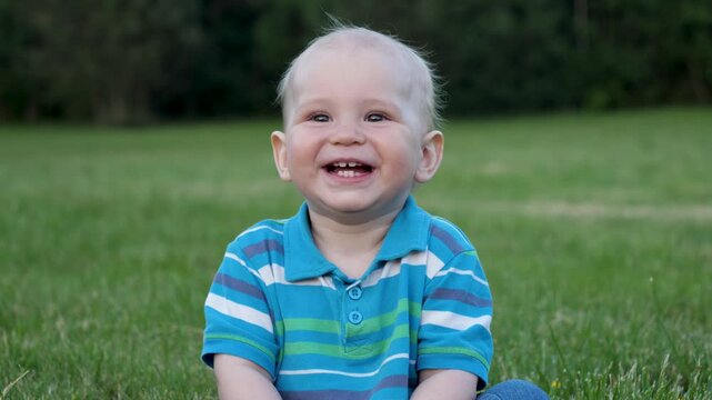 Portrait cheerful toddler with nice big brown eyes sitting on green lawn. Laughing and enjoying a summer day outdoors. Precious moments of childhood joy and innocent happiness in natural park setting