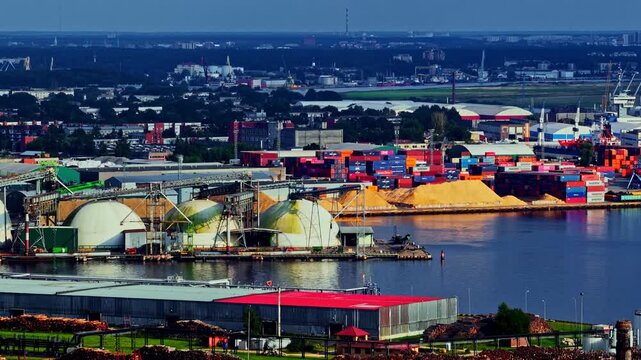 Riga, Latvia - 26 August 2025: Aerial view of the industrial port area featuring large storage domes, colorful shipping containers, and piles of wood chips along the waterfront.