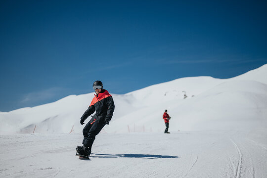 A male snowboarder rides across a groomed snow slope wearing black and red outerwear. The winter scene shows open mountain terrain and a few distant skiers under a clear blue sky.