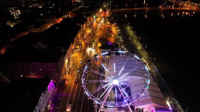 Dynamic aerial view of a sparkling Waterford Christmas waterfront with a towering illuminated observation wheel, decorated facades, and traffic along the river.