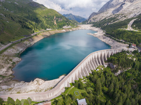 Aerial view of the Fedaia Dam and reservoir in the Dolomites, showing the turquoise water, the concrete dam wall, surrounding mountain slopes, and exposed shoreline in Italy.