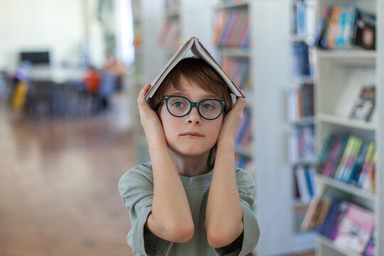 Young boy wearing glasses holds open book over head like roof in library. Back-to-school, preparation for lessons, and pursuit of knowledge. Childhood curiosity,  joy of reading, and love for books.