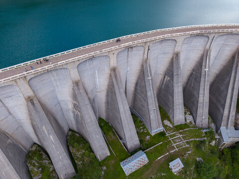 Aerial view of the Fedaia Dam concrete structure in the Dolomites with people walking on the dam crest and green mountain slopes in Italy.
