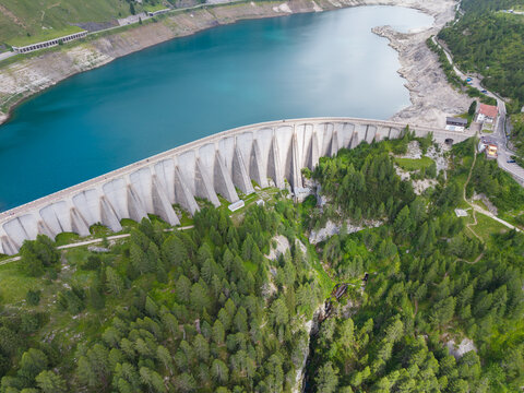 Aerial view of the Fedaia Dam and its reservoir surrounded by lush pine forests and the rugged mountain peaks of the Dolomites in Italy.