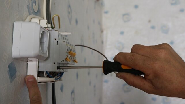 Installing a surface-mounted electrical panel on a wallpapered wall. A technician uses a screwdriver to tighten the contact on a circuit breaker. A white outlet is installed on the left.