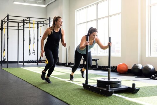 Women pushing sled in gym workout with personal trainer coaching energy