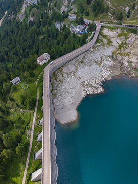Aerial view of the Fedaia Dam and reservoir in the Dolomites, showing exposed rocky shorelines due to low water levels, surrounded by lush alpine forests and mountain slopes in Italy.