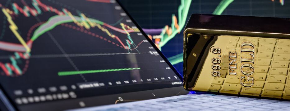 Fine gold bar on laptop keyboard with financial stock market trading chart on screen.