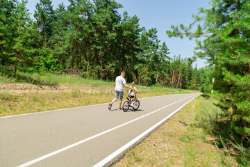 Naklejka premium Child trying the bicycle. Happy father with daughter are having fun in forest at summertime.