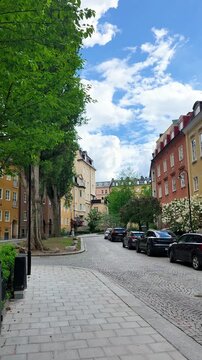 Quiet street on &Ouml;stermalm in Stockholm, Sweden, with colorful historic apartment buildings, parked cars, cobblestone pavement, green trees and cloudy blue sky.