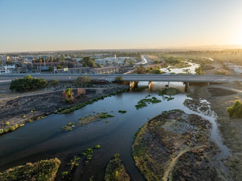 Aerial view of the Kern River flowing under a bridge with surrounding urban infrastructure, dry riverbanks, and golden hour light in Bakersfield, California, United States.