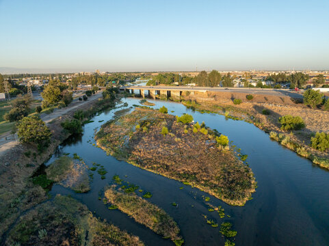 Aerial view of the Kern River flowing through Bakersfield with dry riverbanks, lush vegetation, and a bridge in the background, Bakersfield, California, United States.