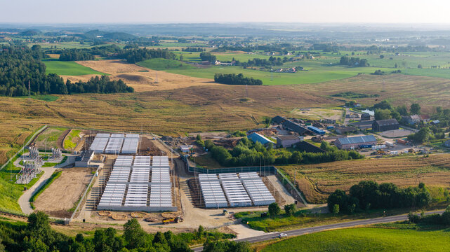 Aerial view of a large scale battery energy storage system with rows of white containers and an electrical substation situated in a rural landscape in Poland.