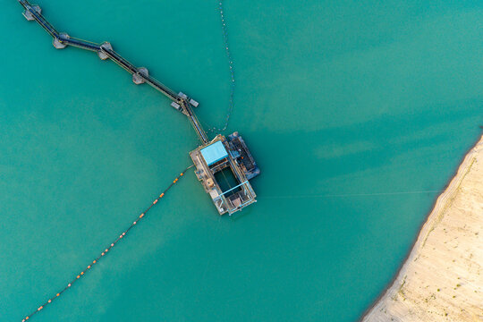Aerial view of a floating and fully automated dredger operating in turquoise water, connected by a long pipeline to the shore, with a sandy bank visible on the right. Poland.