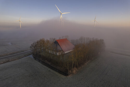 Aerial view of a traditional farm building with a red roof surrounded by trees in a foggy landscape with wind turbines in the background in Cornwerd, Friesland, Netherlands.