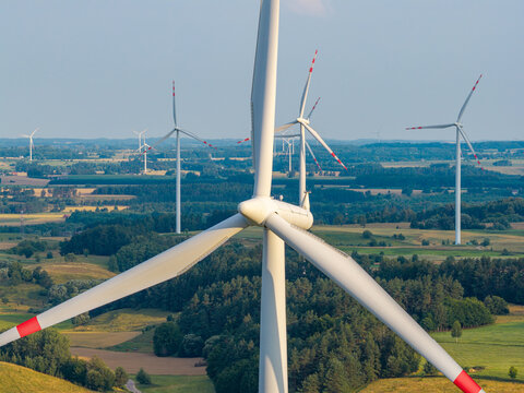 Aerial view of a wind turbine farm with large white blades rotating above a green landscape of rolling hills and forests in Poland.