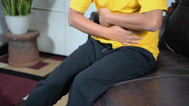 Stomach ache, stomach cramps, heartburn, digestive problems. A young Asian man is holding his stomach and grimacing in pain while sitting on the couch at home.
