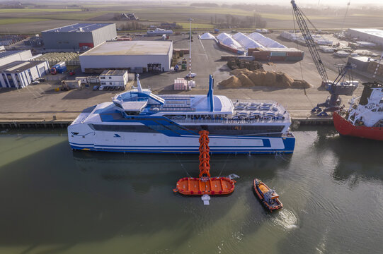 Aerial view of a modern ferry boat undergoing safety training with an orange life raft system deployed in the water at a port in the Netherlands.