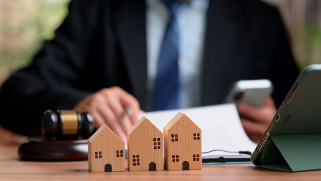 Three small wooden house models sit near auction gavel refocused male attorney reviews real estate contract document holding smartphone device inside courtroom office.