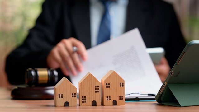 Attorney reviews property contract document behind wooden house models, legal gavel, tablet device, symbolizing real estate law, foreclosure auction, mortgage court case, justice.