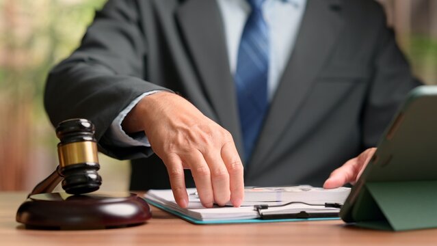 Male lawyer wearing formal suit places hand over cash stack near wooden gavel clipboard digital tablet screen inside office workspace depicting corruption bribery lawsuit settlement legal fee cost.