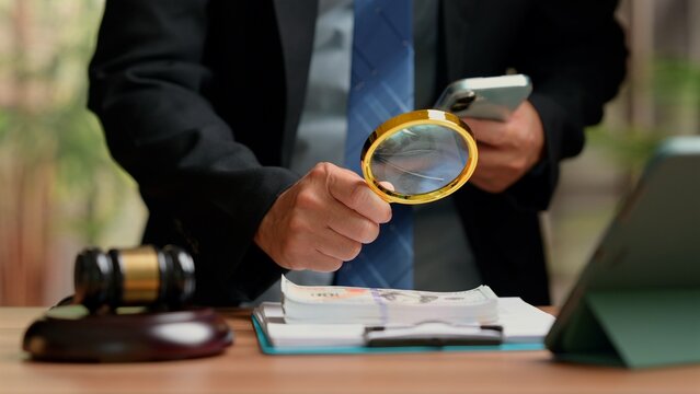 Male forensic investigator wearing suit holding magnifying glass examines dollar banknotes near judge gavel tablet computer office desk conducting financial fraud audit corruption law investigation.