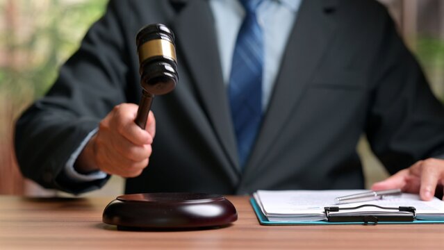Male judge wearing dark suit holding wooden gavel over sound block near legal paper document clipboard inside courtroom office setting before official justice proceeding trial decision.