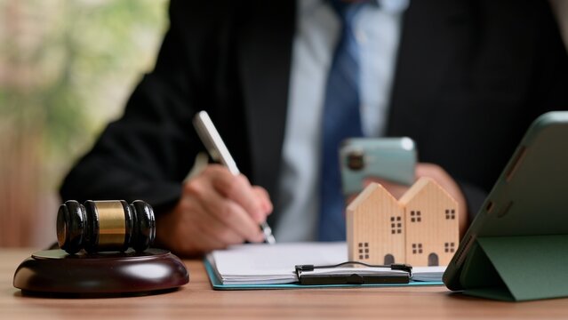 Male corporate attorney signing official real estate contract document inside office, holding smartphone near wooden auction gavel, small wooden house models, digital tablet placed upon office desk.